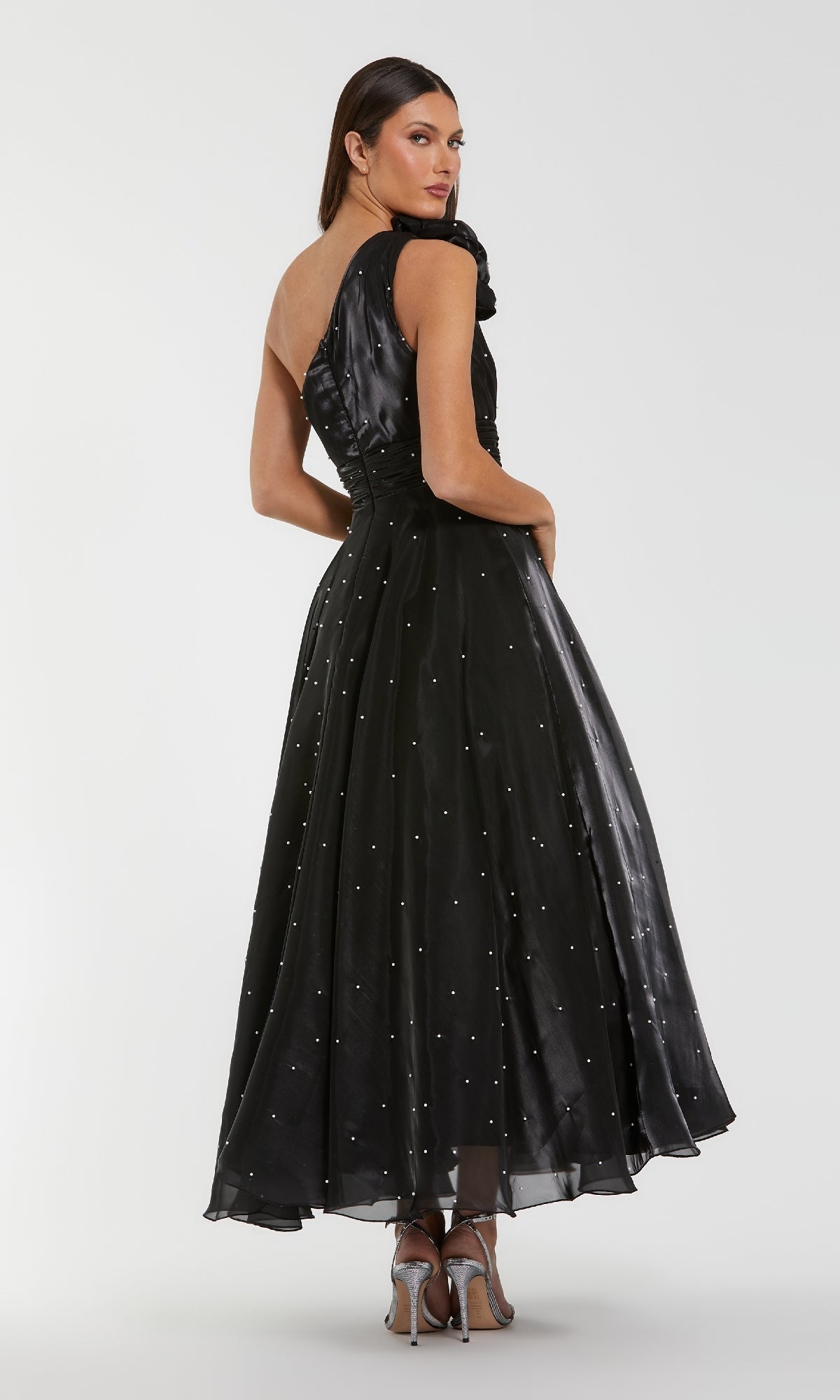 A woman models the Mac Duggal 50078, a one-shoulder black formal dress, featuring pearl beading and subtle embellishments, with silver high heels, posing elegantly against a plain white background.